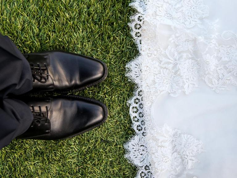 Links schwarze Hose, schwarze Schuhe, rechts der untere Teil eines weißen Kleides. Darunter grüne Wiese.