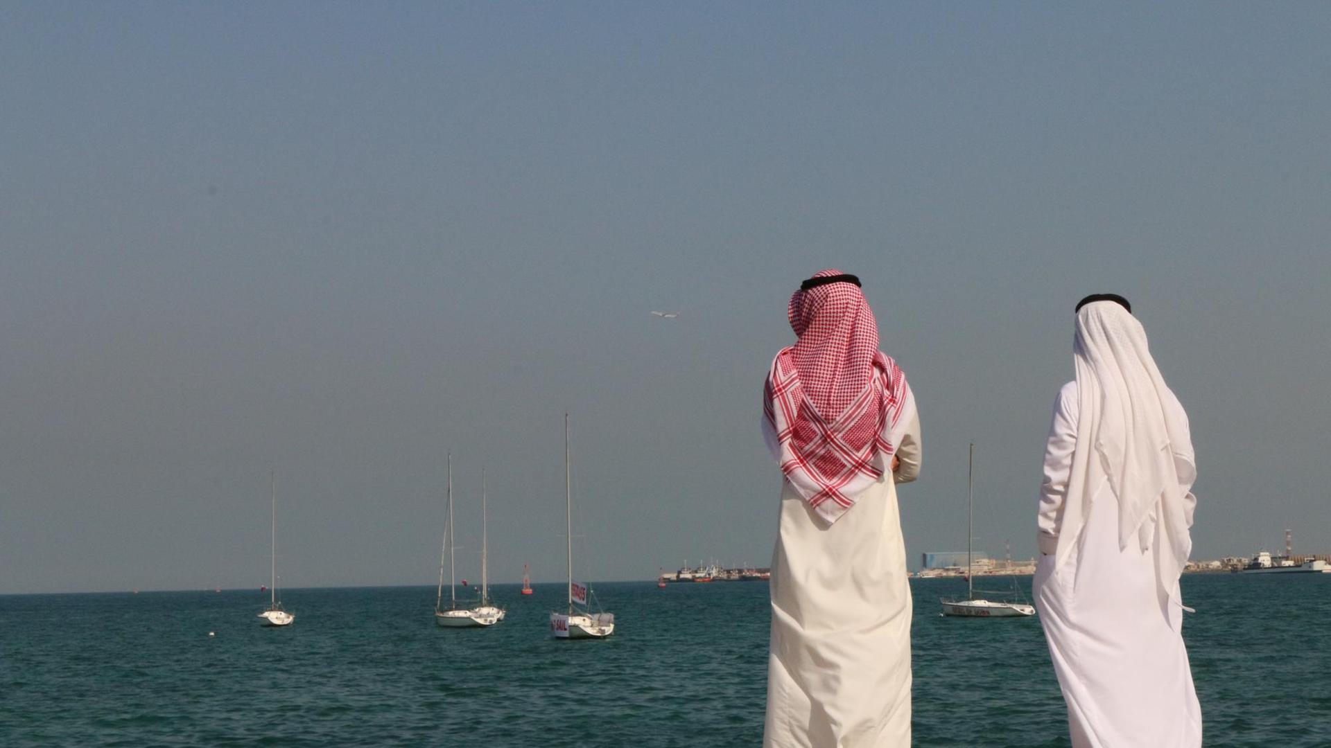 Zwei traditionell gekleidete katarische Bürger stehen an der Uferpromenade der Hauptstadt Doha und blicken auf den Persischen Golf.