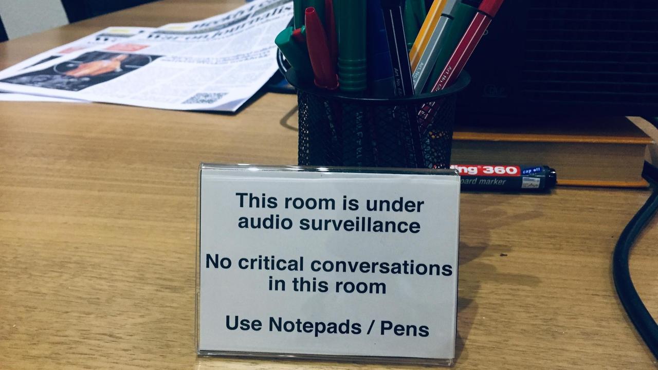 Schreibtisch mit einem Schild, das vor Audio-Überwachung warnt und darum bittet, keine sensiblen Gespräche zu führen. Schreibtisch mit einem Schild, das vor Audio-Überwachung warnt und darum bittet, keine sensiblen Gespräche zu führen.