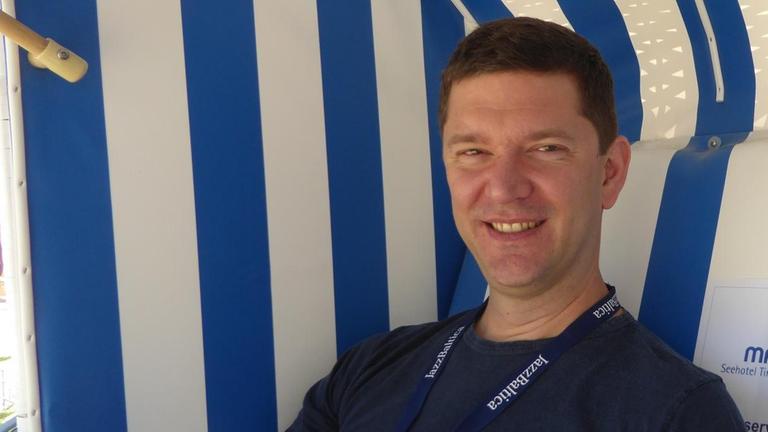 Arne Jansen sitzt lächelnd in einem Strandkorb, um seinen Hals hängt ein Jazzbaltica Festivalpass Arne Jansen sitzt lächelnd in einem Strandkorb, um seinen Hals hängt ein Jazzbaltica Festivalpass