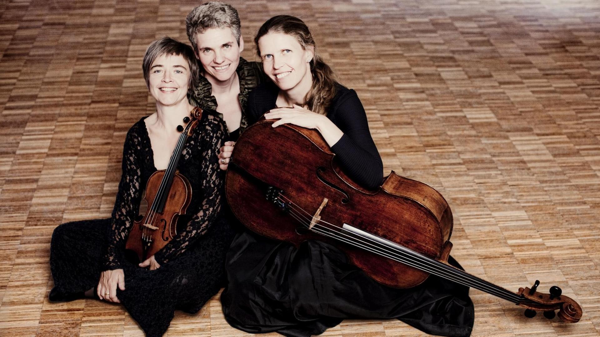 Drei Frauen sitzen schwarz gekleidet auf dem Boden. Eine hält eine Geige in der Hand, eine andere ein Cello. Zusammen bilden sie das Klaviertrio "Trio Vivente".
