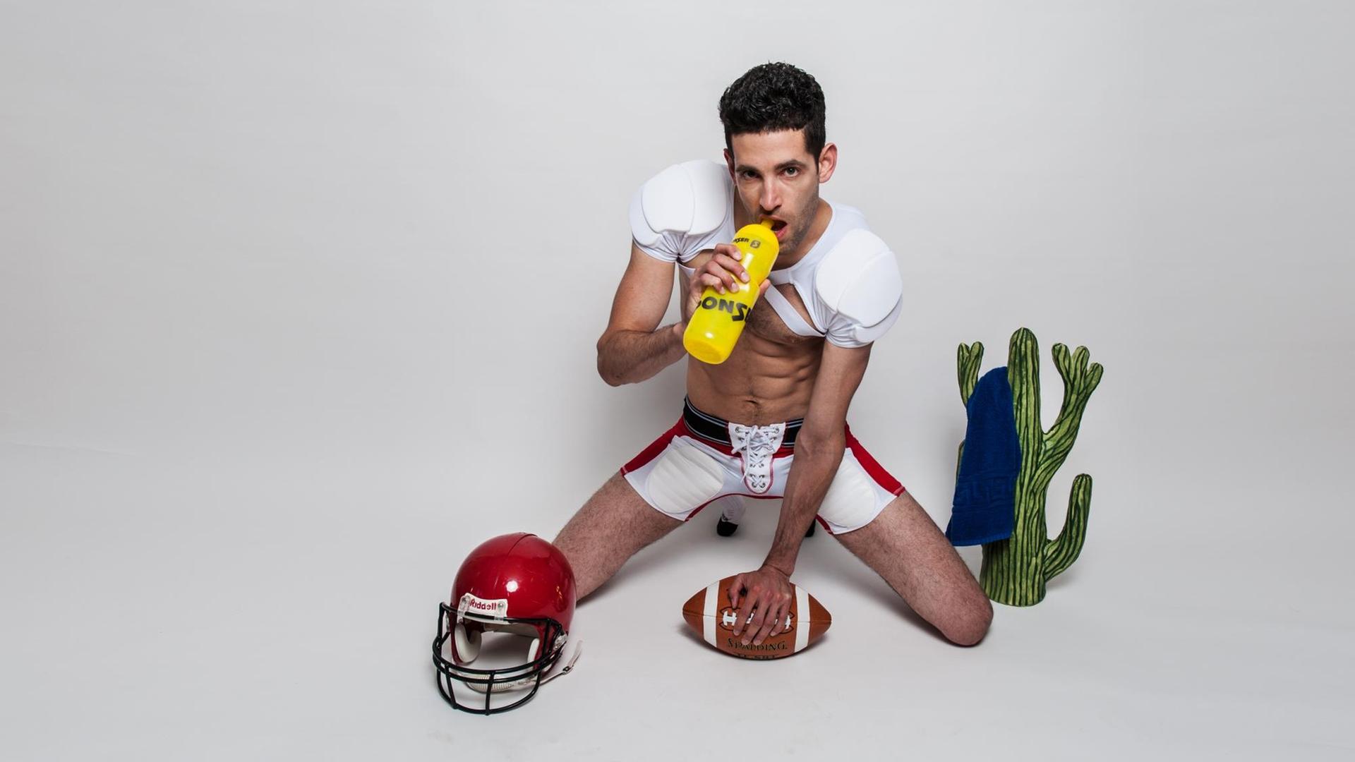Der "Traumboy" in freizügiger Football-Kleidung kniet neben einem Kaktus und nuckelt an einem Sportgetränk (Bild: Maxim-Gorki-Theater Berlin / Raphael Hadad)