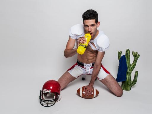 Der "Traumboy" in freizügiger Football-Kleidung kniet neben einem Kaktus und nuckelt an einem Sportgetränk (Bild: Maxim-Gorki-Theater Berlin / Raphael Hadad)