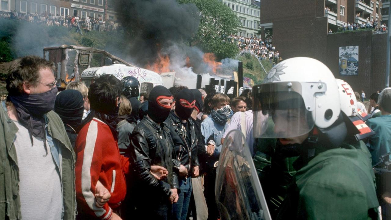 Vermummte Hausbesetzer und Sympathisanten und  weiß behelmte Polizistem  stehen sich am 26.05.1989 vor brennenden Bauwagen in der Hamburger Hafenstraße gegenüber