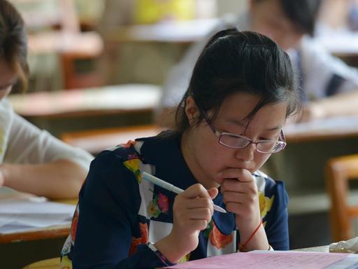 Ein Mädchen sitzt in einem Klassenraum an einem Tisch. Sie ist über ihre Aufgabe gebeugt und denkt nach 