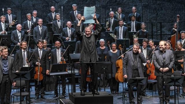 Der Dirigent HK Gruber hält die Partitur zu "Der Prozess" von Gottfried von Einem in die Höhe, vor dem Orchester stehend