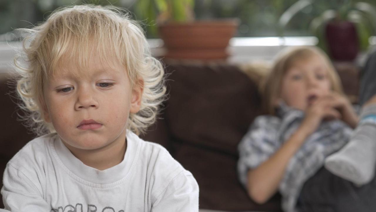 Ein kleiner Junge im Bildvordergrund guckt traurig, im Hintergrund liegt ein größerer Junge auf dem Sofa Ein kleiner Junge im Bildvordergrund guckt traurig, im Hintergrund liegt ein größerer Junge auf dem Sofa