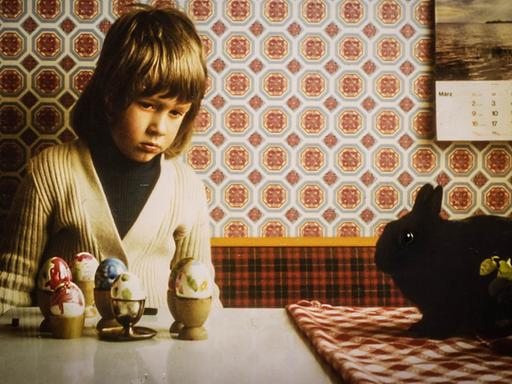 Ein Junge sitzt mit traurigem Gesicht an einem Tisch, auf dem bemalte Ostereier stehen.