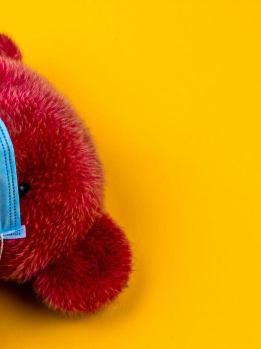 Ein roter Teddybärmit Mundschutz vor gelbem Hintergrund.