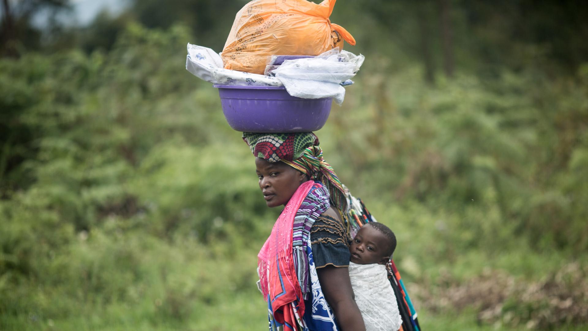 Eine Frau trägt im Distrikt Nyiragongo bei Goma in der Demokratischen Republik Kongo ihr Baby auf dem Rücken und Waren auf dem Kopf. Eine Frau trägt im Distrikt Nyiragongo bei Goma in der Demokratischen Republik Kongo ihr Baby auf dem Rücken und Waren auf dem Kopf.