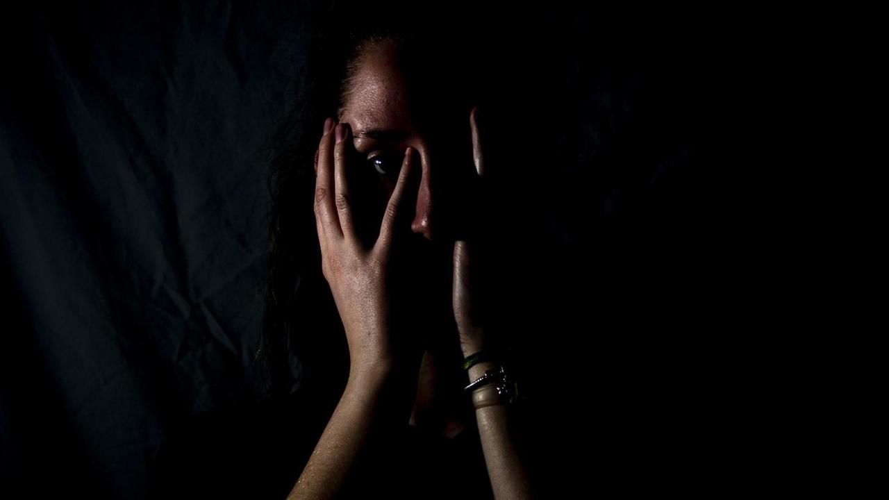 Eine Frau steht im Dunkeln und schlägt erschrocken die Hände vor dem Gesicht zusammen.