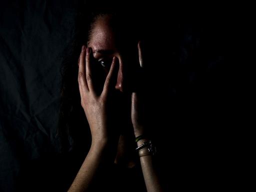 Eine Frau steht im Dunkeln und schlägt erschrocken die Hände vor dem Gesicht zusammen.