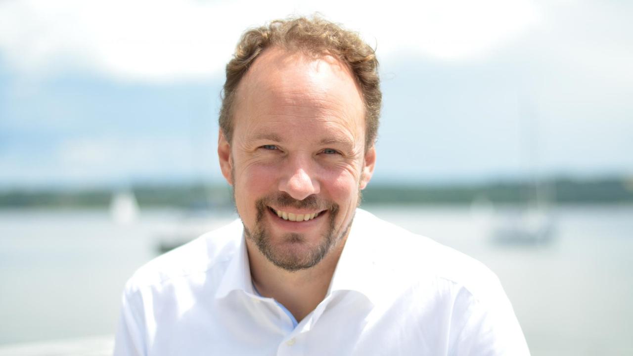 Ein Mann mittleren Alters mit hoher Stirn und Bart sitzt in einem weißen Hemd vor einem See und lacht. Ein Mann mittleren Alters mit hoher Stirn und Bart sitzt in einem weißen Hemd vor einem See und lacht.