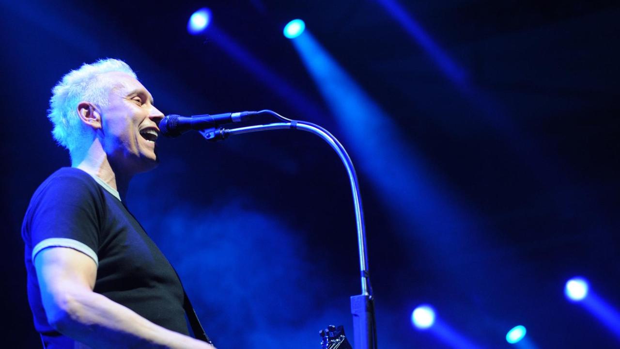 Farin Urlaub ist von der Seite zu sehen, wie er im schwarzen T-Shirt Gitarre spielt und ins Mikrofon singt. Hinter ihm strahlen Scheinwerfer blaues Licht vor schwarzem Hintergrund. Farin Urlaub ist von der Seite zu sehen, wie er im schwarzen T-Shirt Gitarre spielt und ins Mikrofon singt. Hinter ihm strahlen Scheinwerfer blaues Licht vor schwarzem Hintergrund.