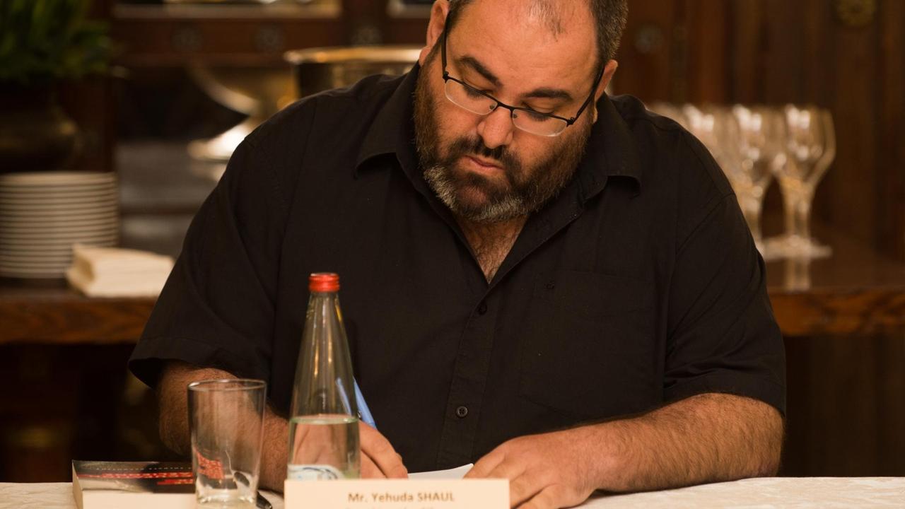 Yehuda Shaul sitzt an einem Konferenztisch und schreibt. Vor ihm ein Namenschild und eine Flasche Wasser. Yehuda Shaul sitzt an einem Konferenztisch und schreibt. Vor ihm ein Namenschild und eine Flasche Wasser.