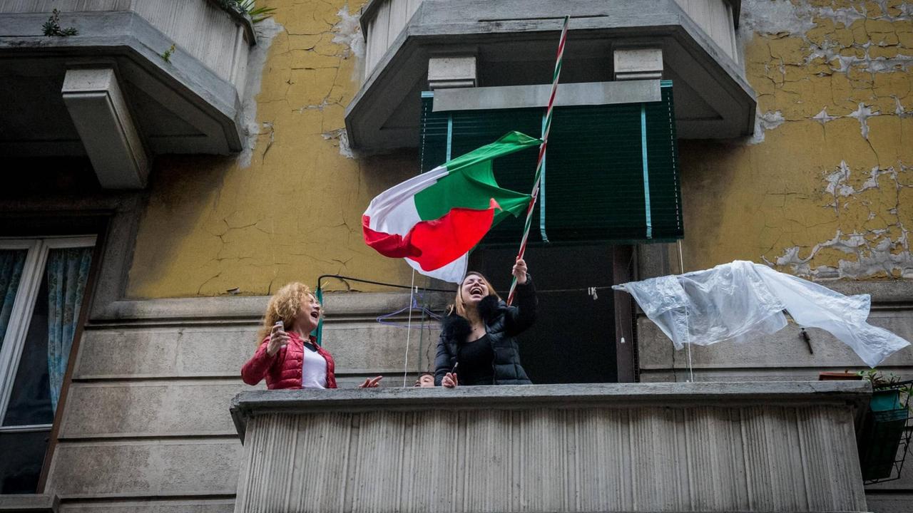 Zwei Frauen stehen singend und lachend auf einem Balkon in Mailand. Eine schwenkt die italienische Nationalflagge. Zwei Frauen stehen singend und lachend auf einem Balkon in Mailand. Eine schwenkt die italienische Nationalflagge.