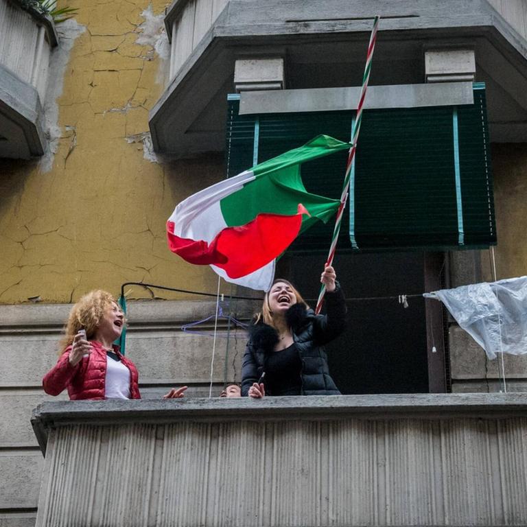 Zwei Frauen stehen singend und lachend auf einem Balkon in Mailand. Eine schwenkt die italienische Nationalflagge. 