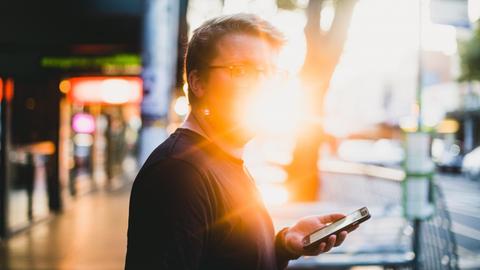 Ein Mann steht am Straßenrand in einer Stadt, ein Smarthone in der Hand. Er schaut Richtung Betrachter, sein Gesicht ist allerdings vom Gegenlicht überblendet.