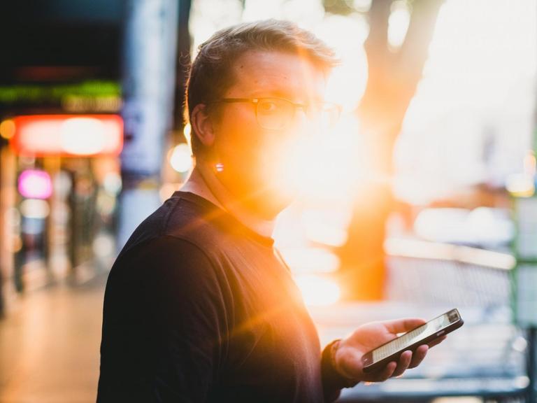 Ein Mann steht am Straßenrand in einer Stadt, ein Smarthone in der Hand. Er schaut Richtung Betrachter, sein Gesicht ist allerdings vom Gegenlicht überblendet.