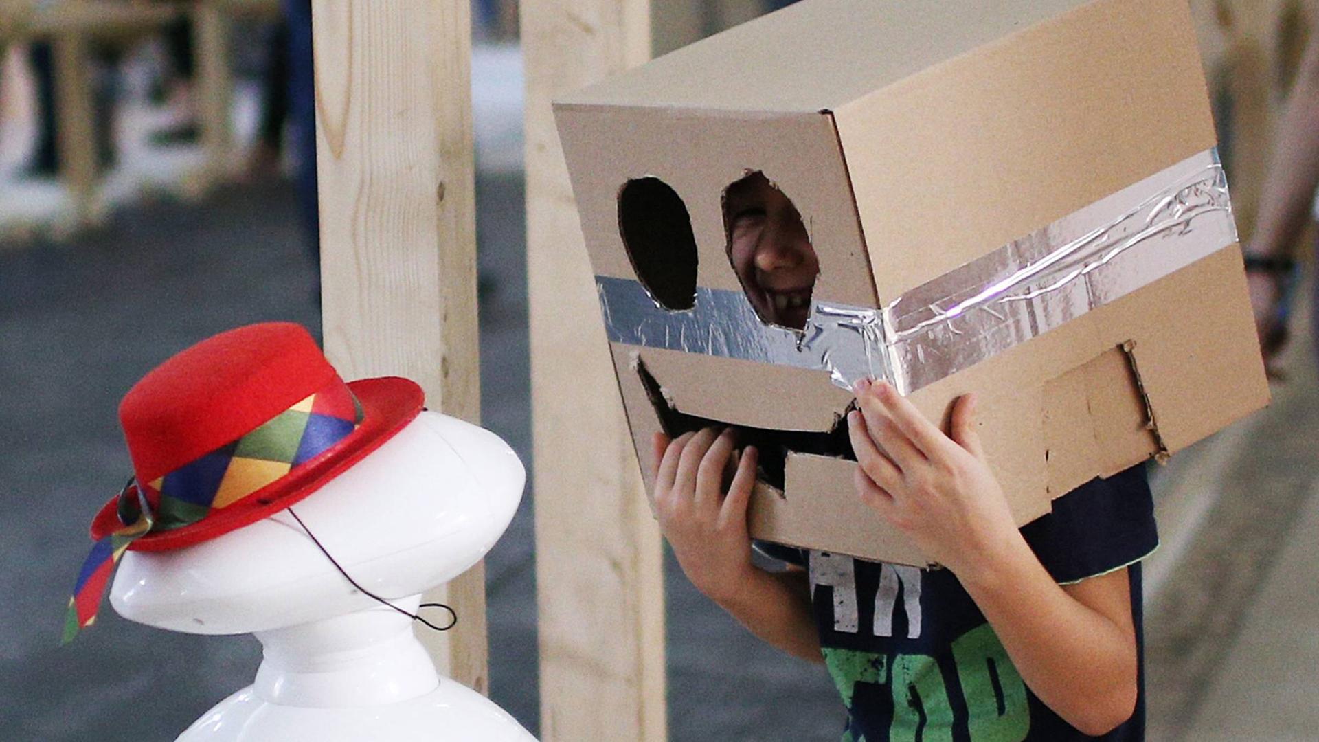 Ein Kind spricht mit einem Computer, während es selbst einen Pappkarton auf dem Kopf trägt, in dessen Front ein Gesicht geschnitten ist.