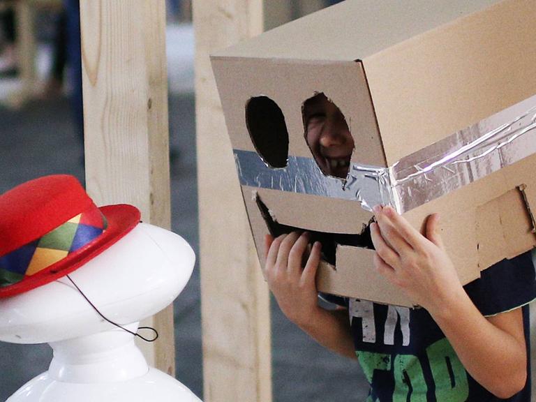 Ein Kind spricht mit einem Computer, während es selbst einen Pappkarton auf dem Kopf trägt, in dessen Front ein Gesicht geschnitten ist.