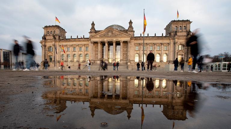 Durch eine Langzeitbelichtung verschwimmen die vorbeigehenden Personen vor dem Bundestag, welcher sich in einer großen Pfütze spiegelt. Durch eine Langzeitbelichtung verschwimmen die vorbeigehenden Personen vor dem Bundestag, welcher sich in einer großen Pfütze spiegelt.