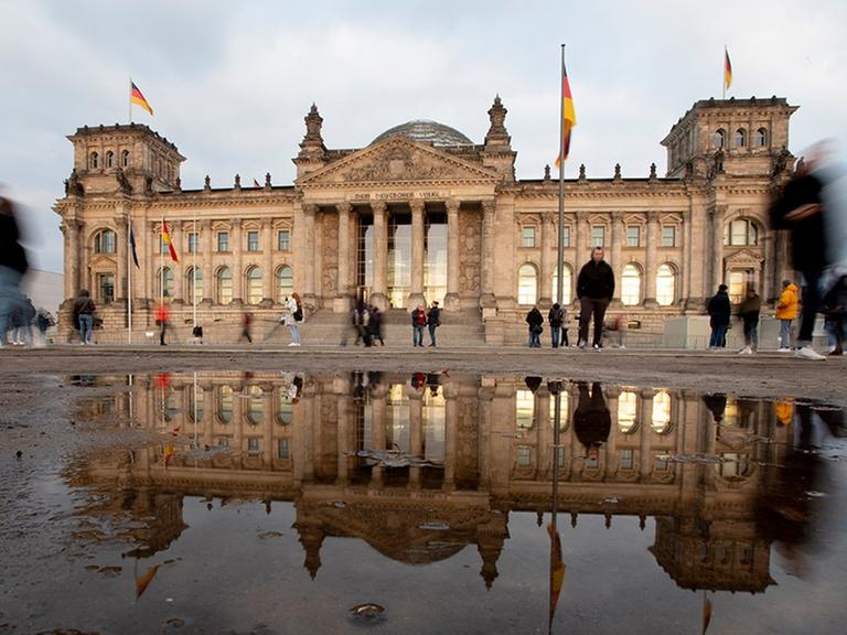 Durch eine Langzeitbelichtung verschwimmen die vorbeigehenden Personen vor dem Bundestag, welcher sich in einer großen Pfütze spiegelt.