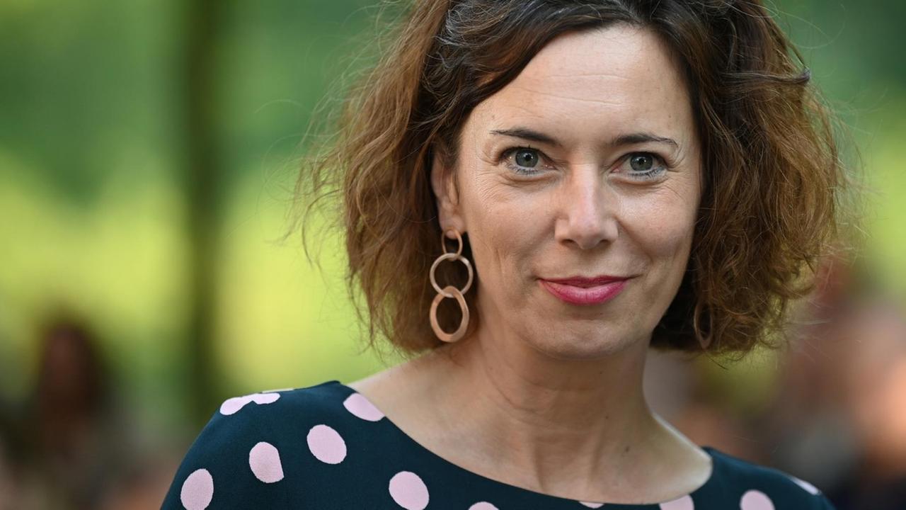 Die österreichische Schriftstellerin Eva Menasse lächelt in die Kamera. Sie hat eine Sonnenbrille ins Haar geschoben, trägt Ohrringe und ein blaues Kleid mit weißen Punkten. Die österreichische Schriftstellerin Eva Menasse lächelt in die Kamera. Sie hat eine Sonnenbrille ins Haar geschoben, trägt Ohrringe und ein blaues Kleid mit weißen Punkten.