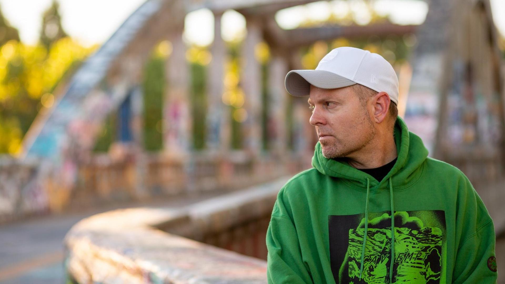 DJ Shadow zur Seite blickend, mit grünem Hoodie und weißem Basecap; im Hintergrund eine Brücke