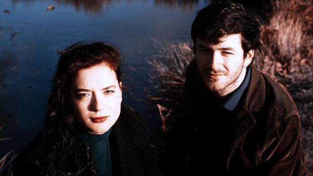 Eine Frau (links) und ein Mann, beide mit dunklen Haaren, stehen am Ufer eines Teichs und schauen direkt in die Kamera, die sie von schräg oben aufnimmt.