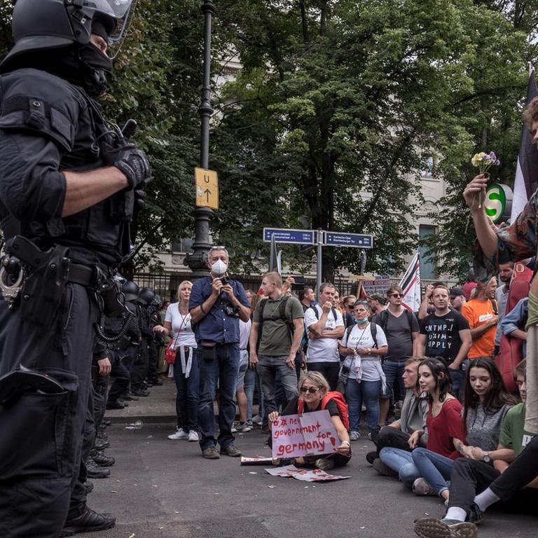 Eine Straßenszene während der Demonstration gegen die Corona-Maßnahmen am 29. August in Berlin. Darauf zu sehen ist ein Polizist, der diversen Demonstranten gegenüber steht. Einer davon hält in hippiesker Kleidung eine Blume in der Hand. Hinter ihm sind schwarz-weiß-rote Reichskriegsflagge aus Zeiten des Deutschen Kaiserreich zu sehen.