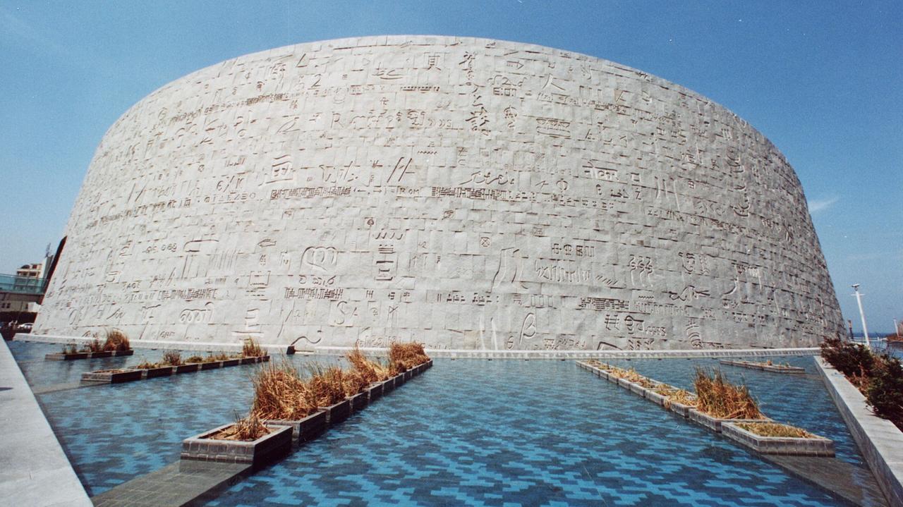 Ein Blick auf die Außenmauer der Bibliothek von Alexandria: Ein runder Bau mit ägyptischen Schriftzeichen auf der grauen Betonwand. Davor befinden sich Wasserflächen.