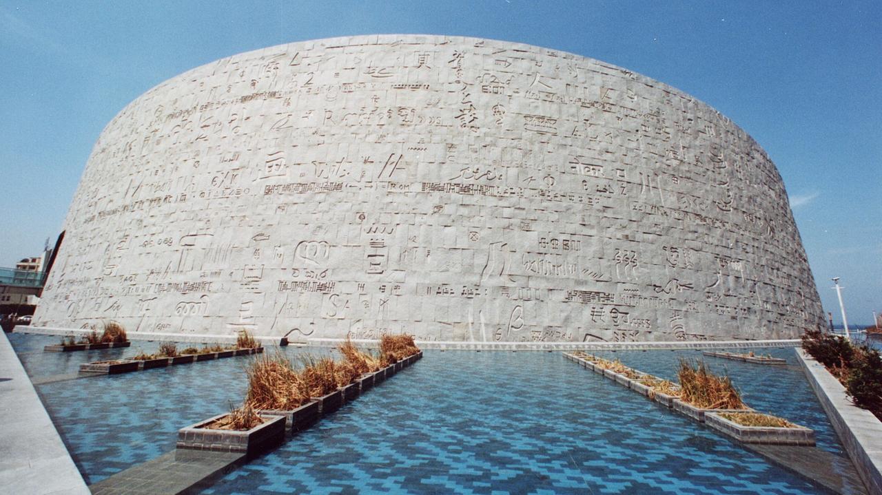 Ein Blick auf die Außenmauer der Bibliothek von Alexandria: Ein runder Bau mit ägyptischen Schriftzeichen auf der grauen Betonwand. Davor befinden sich Wasserflächen. Ein Blick auf die Außenmauer der Bibliothek von Alexandria: Ein runder Bau mit ägyptischen Schriftzeichen auf der grauen Betonwand. Davor befinden sich Wasserflächen.