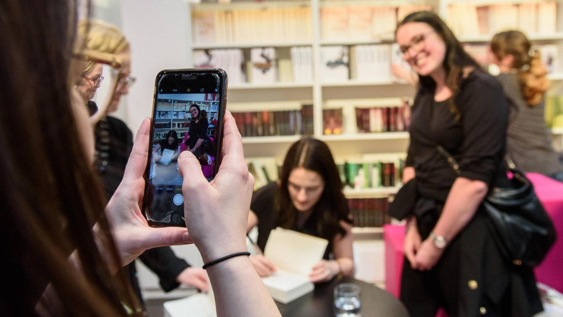Eine junge Frau fotografiert mit einem Smartphone eine Signierstunde: Autorin Mona Kasten signiert, ein weiblicher Fan lächelt in die Kamera. Eine junge Frau fotografiert mit einem Smartphone eine Signierstunde: Autorin Mona Kasten signiert, ein weiblicher Fan lächelt in die Kamera.