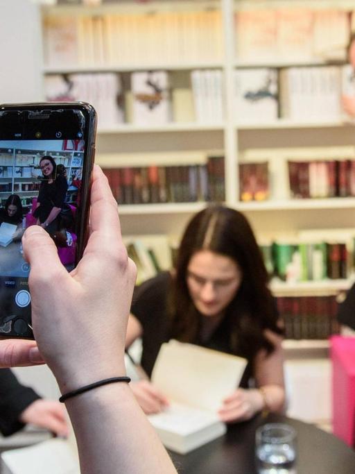 Eine junge Frau fotografiert mit einem Smartphone eine Signierstunde: Autorin Mona Kasten signiert, ein weiblicher Fan lächelt in die Kamera.