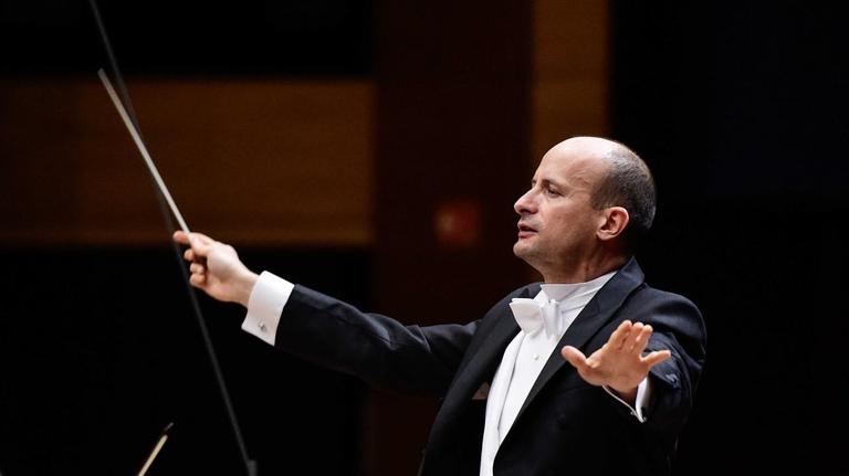 Seitlich ist ein Dirigent mit ausgebreiteten Armen und erhobenem Kopf zu sehen, der vor dem Orchester steht und dirigiert. Seitlich ist ein Dirigent mit ausgebreiteten Armen und erhobenem Kopf zu sehen, der vor dem Orchester steht und dirigiert.