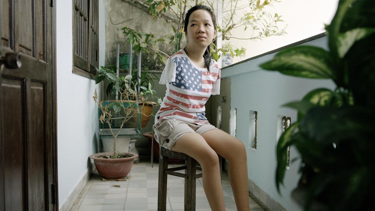 Eine junge Frau sitzt auf einem Hocker auf einem Balkon. Sie hat keine Arme und trägt ein T-Shirt mit aufgedruckter US-Flagge. Eine junge Frau sitzt auf einem Hocker auf einem Balkon. Sie hat keine Arme und trägt ein T-Shirt mit aufgedruckter US-Flagge.