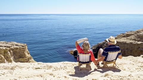 Ein Paar sitzt in Liegestühlen am Mittelmeer in der Sonne, die Frau trägt eine Armschiene.