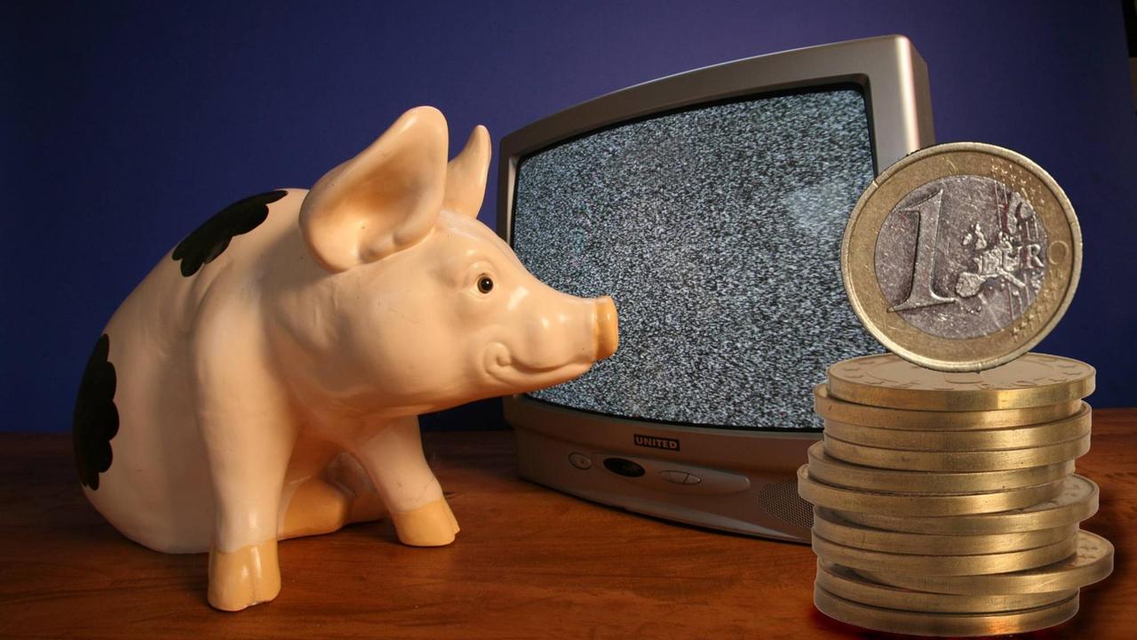 Ein Plastik-Schweinchen starrt auf den leeren Bildschirm eines Fernsehers, vor dem Ein-Euro-Münzen gestapelt sind