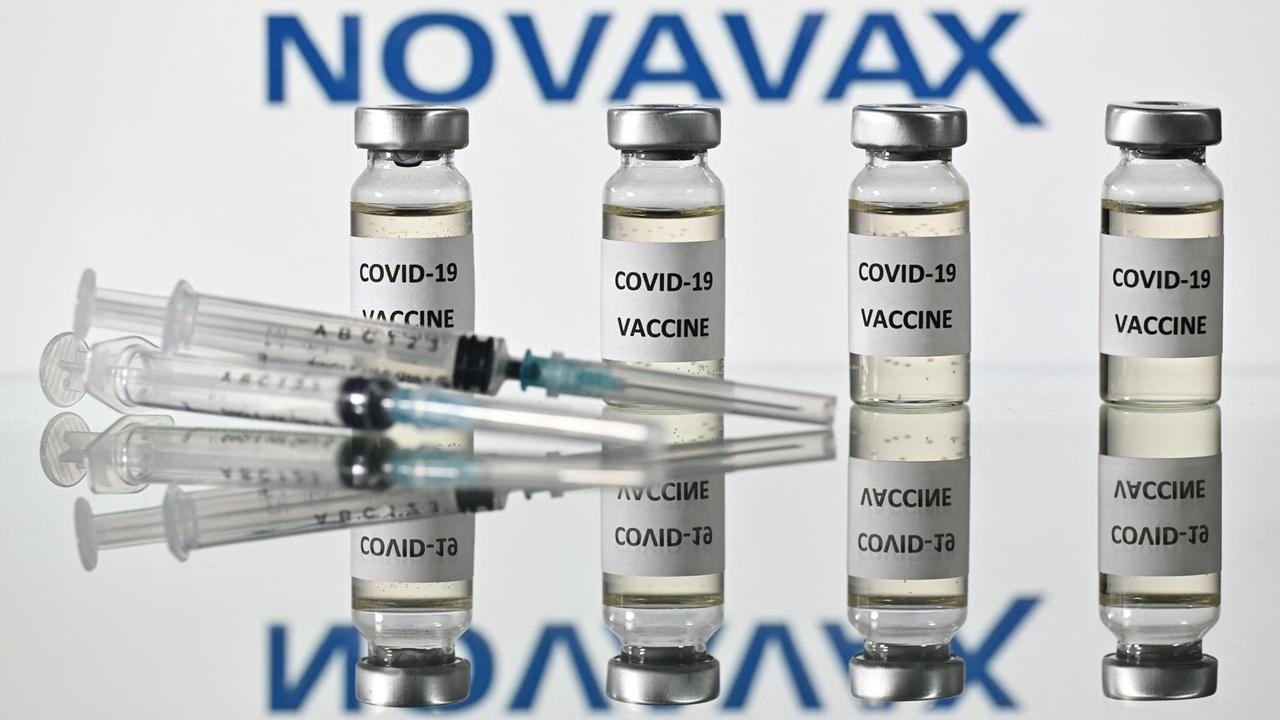 Zwei Spritzen liegen vor vier Ampullen gefüllt mit dem Impfstoff des Herstellers Novavax. Sein Firmenlogo prangert im Hintergrund. 