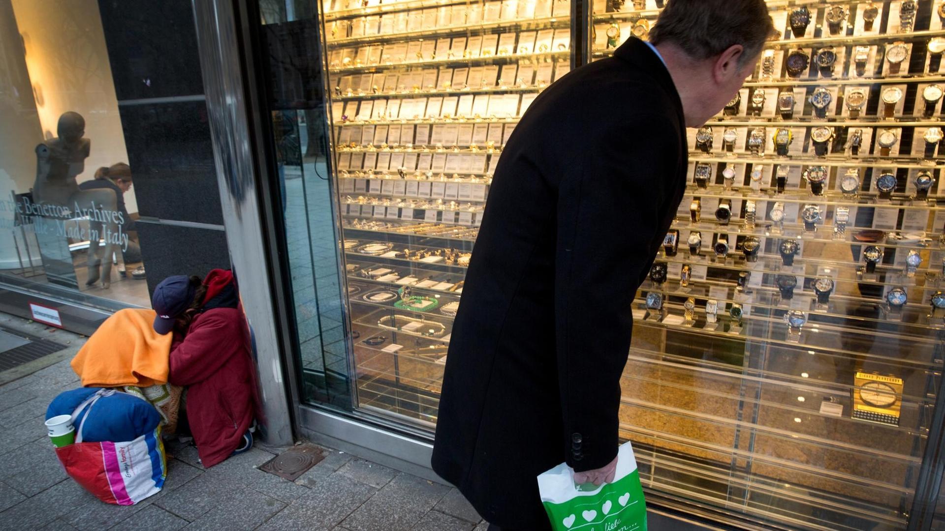 Ein Mann schaut am in Düsseldorf auf der Königsallee in ein Schaufenster mit Luxusuhren, während neben ihm ein Bettler um Geld bittet. Ein Mann schaut am in Düsseldorf auf der Königsallee in ein Schaufenster mit Luxusuhren, während neben ihm ein Bettler um Geld bittet.