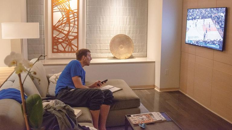 Dirk Nowitzki sitzt auf der Couch in einem Hotelzimmer und schaut ein NBA-Spiel auf einem Fernseher. Dirk Nowitzki sitzt auf der Couch in einem Hotelzimmer und schaut ein NBA-Spiel auf einem Fernseher.