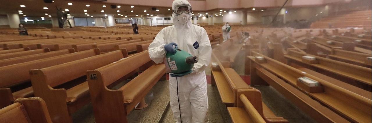 Eine Person in Schutzkleidung versprüht aus einem Kanister Desinfektionsmittel zwischen den Kirchenbänken.