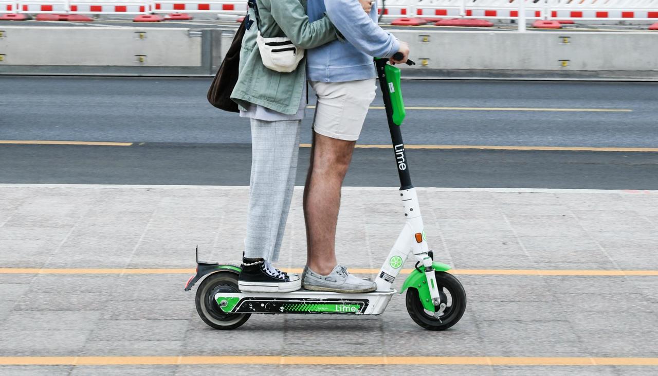 Seitenansicht von zwei Personen, die gemeinsam auf einem E-Tretroller fahren. Seitenansicht von zwei Personen, die gemeinsam auf einem E-Tretroller fahren.