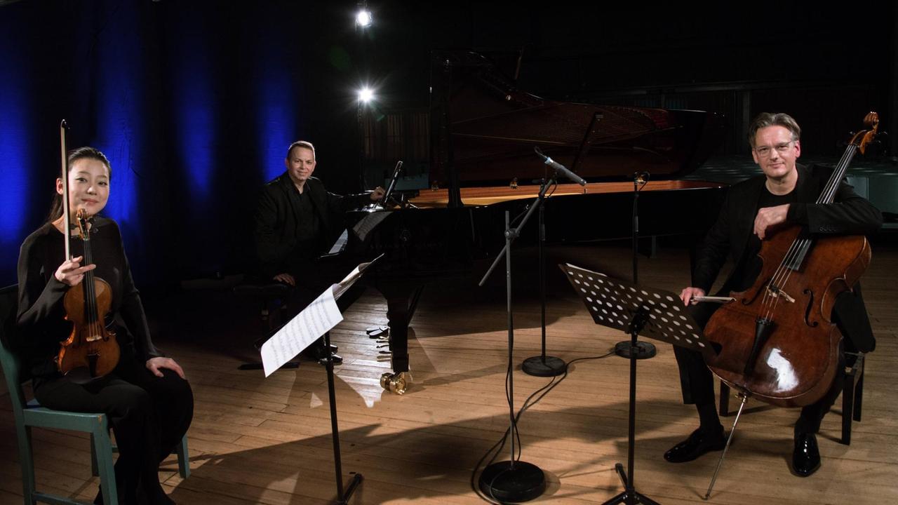 Die drei Musiker sitzen in einem abgedunkelten Raum, nur ihr Kammermusikbereich ist beleuchtet, und lächeln in die Kamera. Die drei Musiker sitzen in einem abgedunkelten Raum, nur ihr Kammermusikbereich ist beleuchtet, und lächeln in die Kamera.