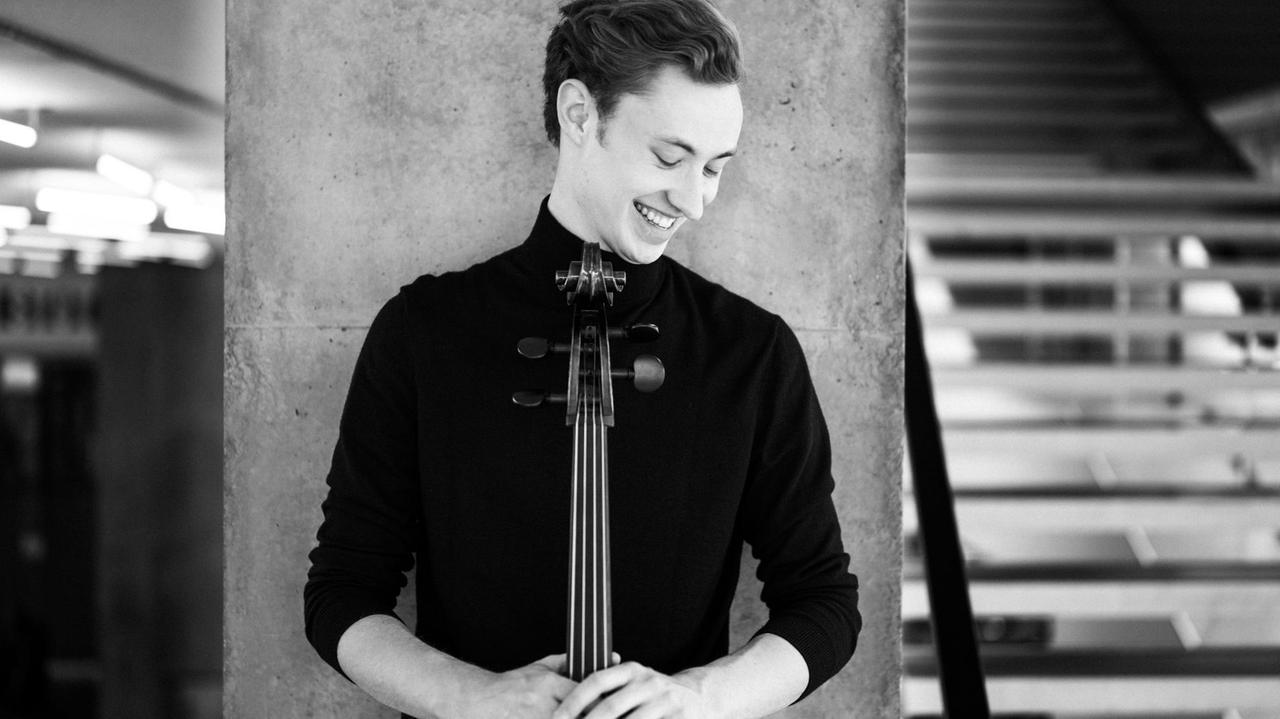 Der Cellist steht mit seinem Instrument vor einer grauen Betonwand in einem größeren Treppenhaus. Der Cellist steht mit seinem Instrument vor einer grauen Betonwand in einem größeren Treppenhaus.