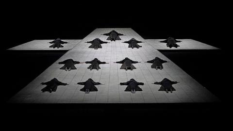 13 dunkel gekleidete Nonnen liegen auf einem riesigen Bühnen-Holzboden in Form eines Kreuzes.