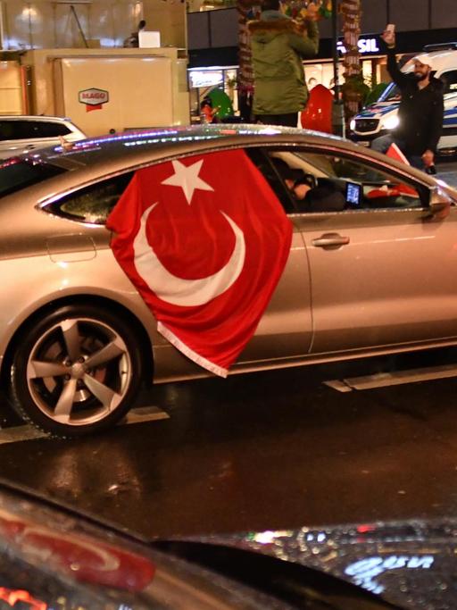Sie sehen ein silbernes Auto mit einer Türkeifahne in Berlin. Es ist Nacht.