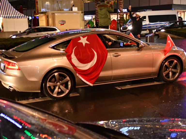 Sie sehen ein silbernes Auto mit einer Türkeifahne in Berlin. Es ist Nacht.