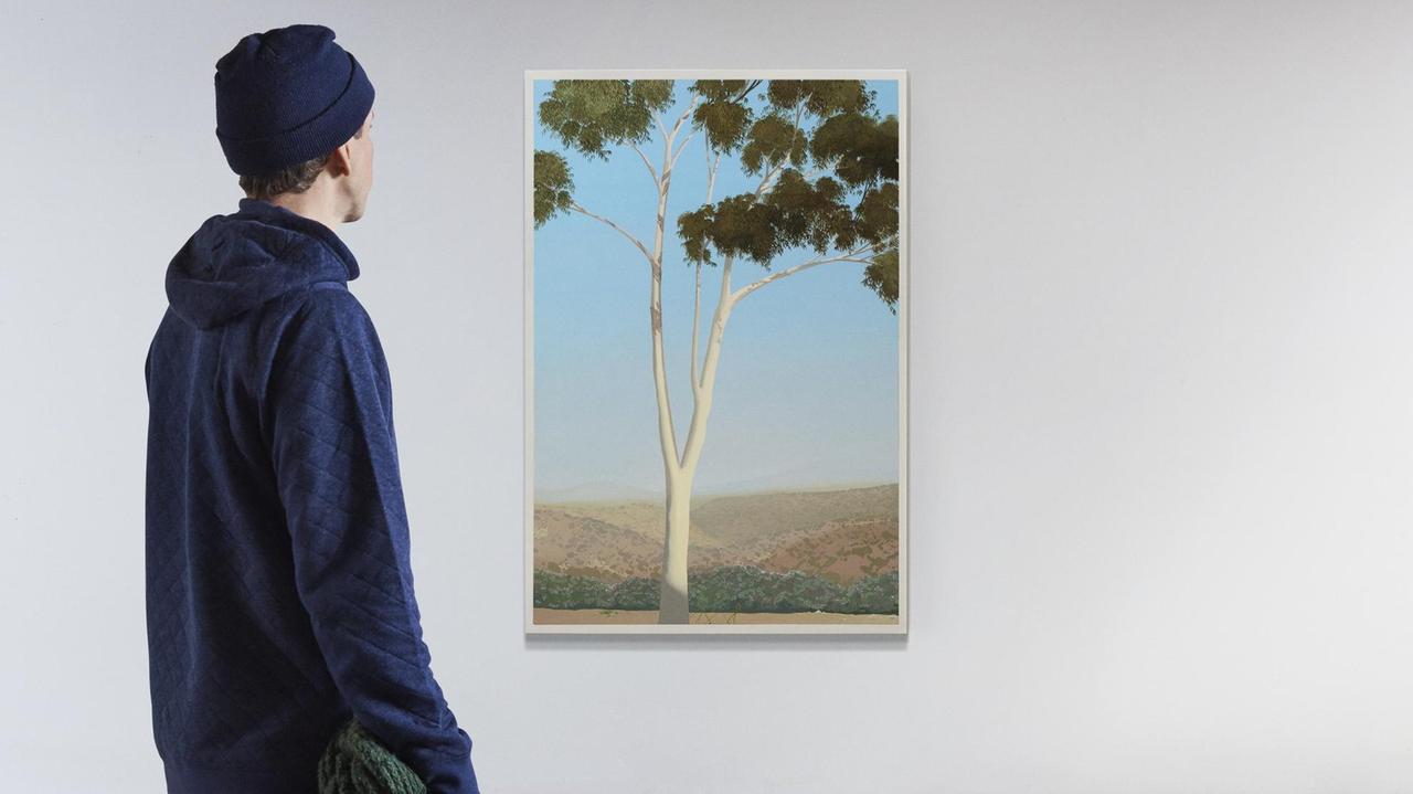 Ein Mann blickt auf ein Bild eines Baumes, das an einer leeren, weißen Wand hängt. Man sieht ihn von hintern. Er trägt eine dunkelblaue Mütze und einen dunkelblauen Pullover. Ein Mann blickt auf ein Bild eines Baumes, das an einer leeren, weißen Wand hängt. Man sieht ihn von hintern. Er trägt eine dunkelblaue Mütze und einen dunkelblauen Pullover.